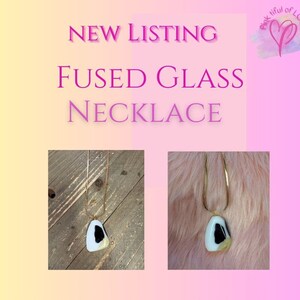 May include: A fused glass necklace with a white, black, and yellow triangular pendant on a gold chain. The necklace is photographed on a wooden surface and a fuzzy white surface.