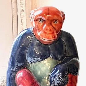 May include: A ceramic monkey figurine with a red face and legs, a blue body, and a green stomach. The monkey is in a seated position with its legs crossed and hands resting on its knees. The figurine has a glossy finish.
