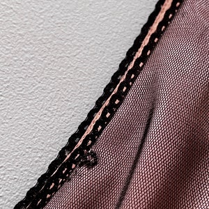 May include: Close-up of a sheer pink fabric with a black lace trim. The fabric has a fine mesh pattern.