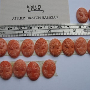 Puede incluir: Una colección de 14 colgantes de camafeo de forma ovalada que presentan un perfil de mujer en relieve. Los camafeos están hechos de un material rosa claro y están dispuestos en dos filas sobre una superficie blanca. El texto "ATELIER HRATCH BABIKIAN" es visible en un trozo de papel en el fondo.