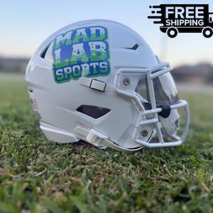 May include: A white football helmet featuring a green and blue logo on the side that reads MAD LAB SPORTS. The helmet includes a white face mask, a clear visor, and a small American flag decal on the rear side, presented in a glossy finish.