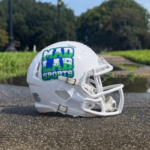 May include: A white football helmet featuring a white face mask and a green and blue graphic decal on the side that reads MAD LAB SPORTS. The helmet has a glossy finish and includes small ventilation holes along the side and back shell.
