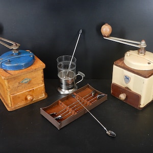 May include: Two vintage wooden coffee grinders with a blue and white painted top, a brown wooden drawer, and a wooden handle. A silver metal cup with a glass insert and a silver metal stirrer. A brown leather case with six silver metal spoons.