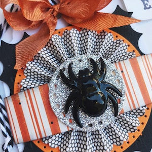 May include: A close-up of a Halloween-themed craft project featuring a black spider with silver glitter on a white and black polka dot background. The spider is surrounded by a white and orange striped ribbon and a black and white polka dot paper fan.