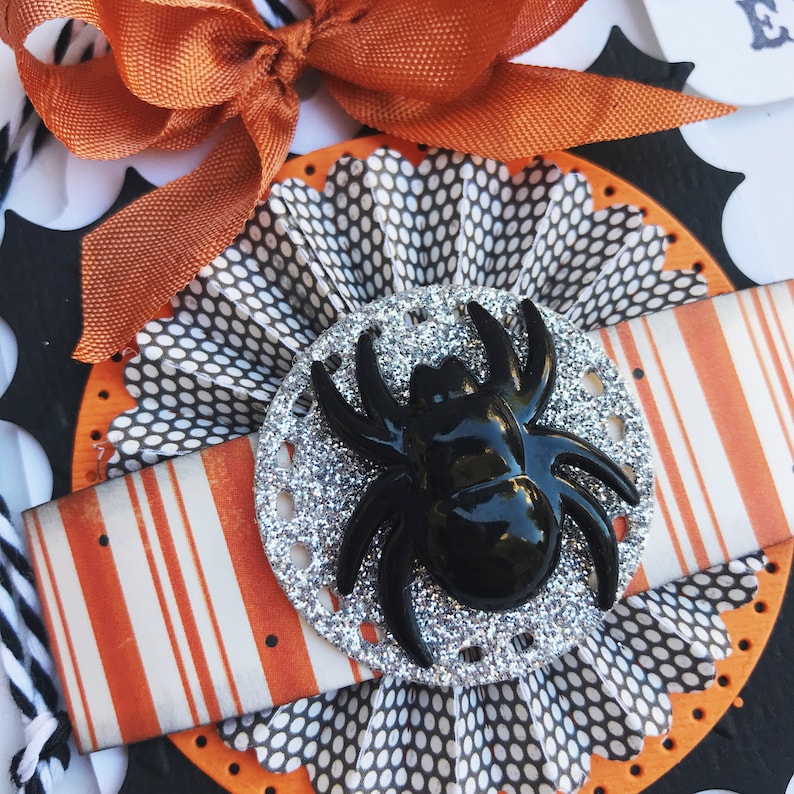 May include: A close-up of a Halloween-themed craft project featuring a black spider with silver glitter on a white and black polka dot background. The spider is surrounded by a white and orange striped ribbon and a black and white polka dot paper fan.