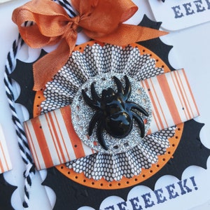 May include: A black and orange Halloween card with a spider on a silver glitter circle. The card has a scalloped edge and says "Eeek!" in white letters.