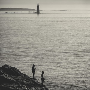May include: Two people silhouetted against a rocky shore, fishing with rods in a body of water. A lighthouse stands in the distance on a small island.