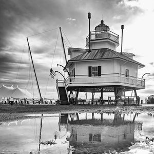 May include: Black and white photograph of a lighthouse with a raised structure, a small balcony, and a cupola. The lighthouse is reflected in a puddle of water in the foreground. An American flag is visible.