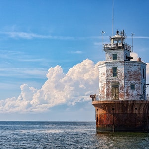 Könnte beinhalten: Ein verwitterter Leuchtturm steht im Meer unter blauem Himmel mit weißen Wolken. Der Leuchtturm ist weiß mit roten Backsteinakzenten und einem rostbraunen Sockel. Er hat einen kleinen Turm mit Antennen.