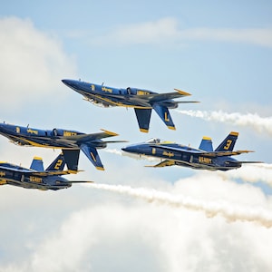 Könnte beinhalten: Vier blaue und gelbe Kampfjets fliegen in Formation und hinterlassen weiße Rauchspuren vor einem blauen Himmel mit weißen Wolken.