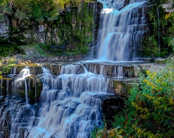 Peut inclure: Une cascade pittoresque dévale des formations rocheuses en couches, entourée d'une végétation luxuriante et d'arbres aux couleurs automnales vives. L'eau en mouvement est floutée, créant une sensation de mouvement et de tranquillité.