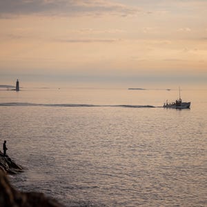 May include: A silhouette of two people standing on a rocky shore, looking out at a boat on the water. The boat is in the distance, and the sky is a soft pink and orange.