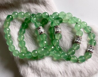 Può includere: Quattro braccialetti in giada verde con accenti argentati e cristalli trasparenti. I braccialetti sono impilati uno sopra l'altro e sono su uno sfondo di pelliccia sintetica bianca.