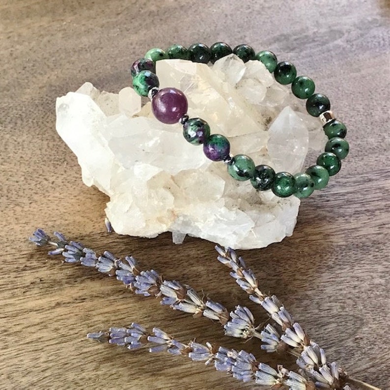 May include: A green and purple gemstone bracelet with a single larger purple gemstone bead. The bracelet is resting on a white crystal cluster.