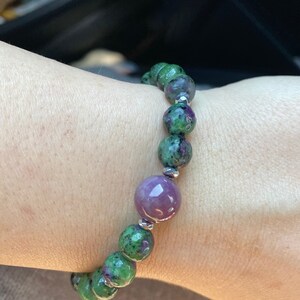 May include: A green and purple gemstone bracelet with silver beads. The bracelet is made of round beads and has a larger, round, purple gemstone in the center.