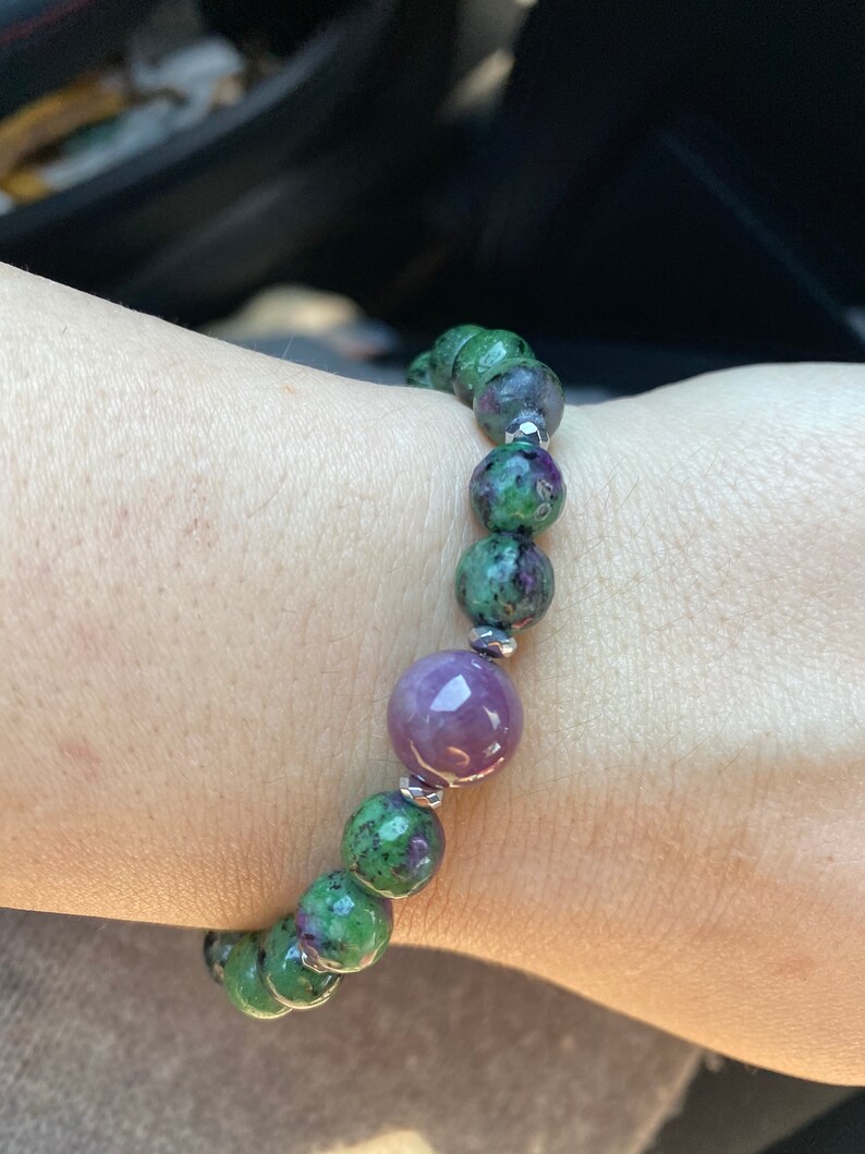 May include: A green and purple gemstone bracelet with silver beads. The bracelet is made of round beads and has a larger, round, purple gemstone in the center.
