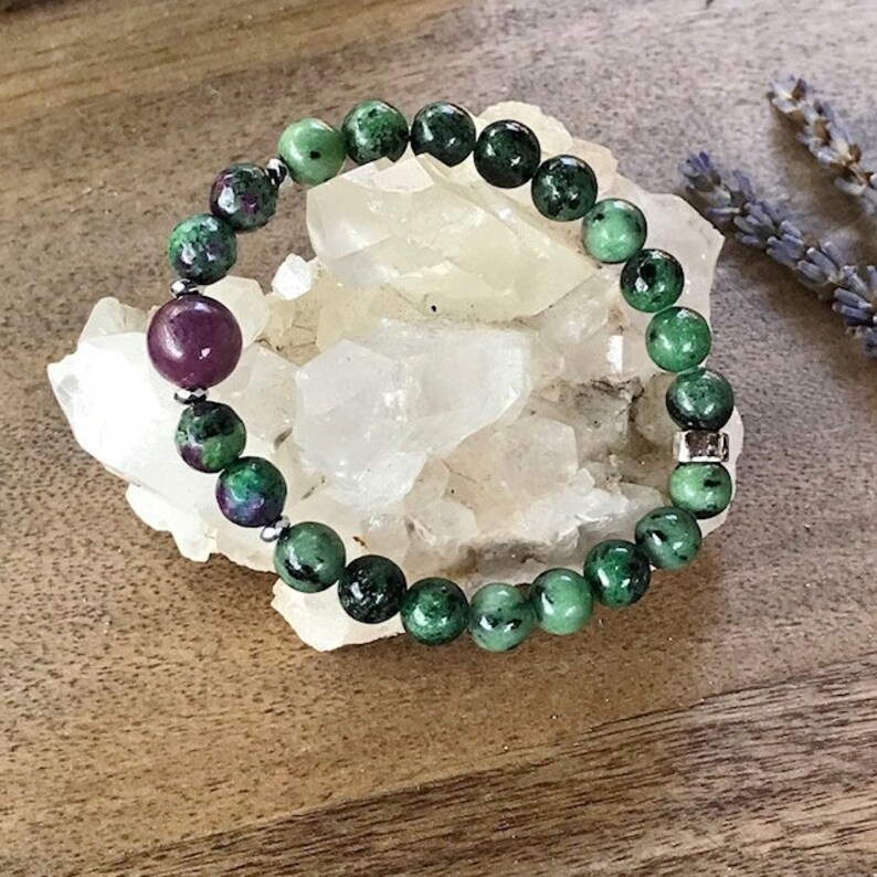 May include: A green and pink gemstone bracelet with silver beads. The bracelet is made of round beads and is lying on a white crystal.