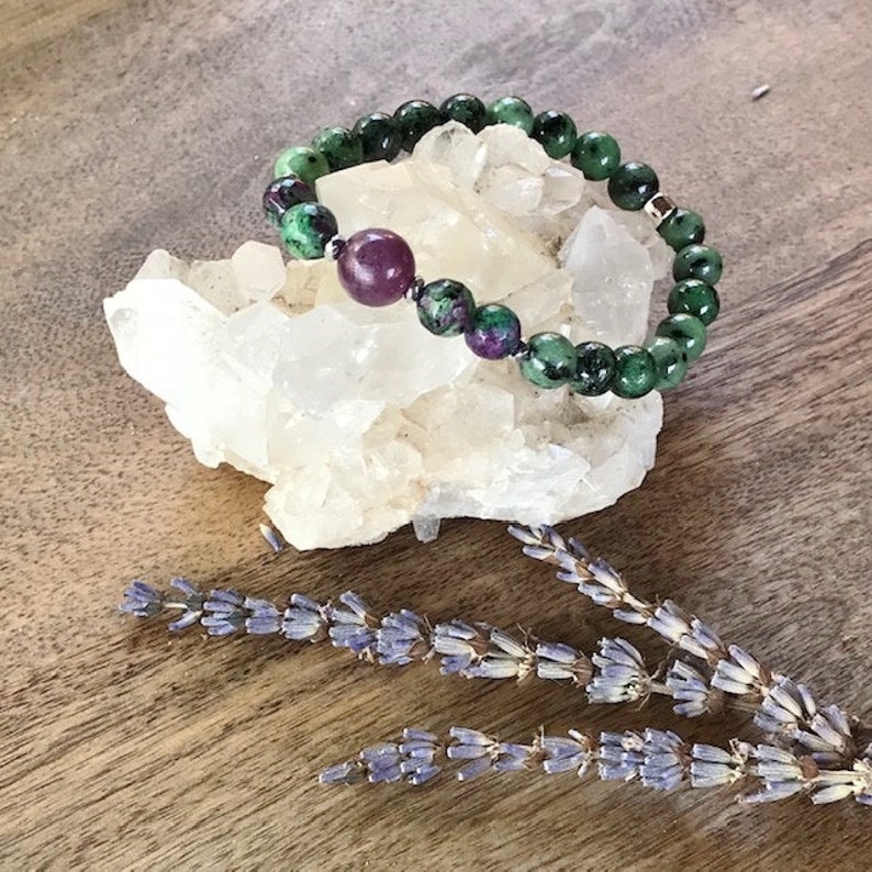 May include: A green and purple gemstone bracelet with a silver clasp. The bracelet is on a white crystal cluster.