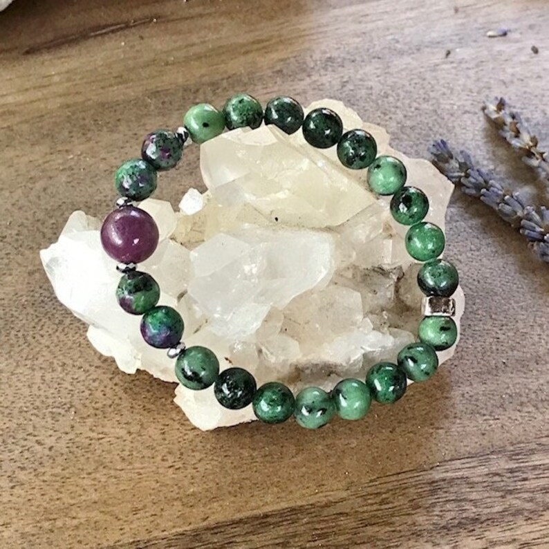 May include: A green and pink gemstone bracelet with silver beads. The bracelet is made of round beads and is lying on a white rock.