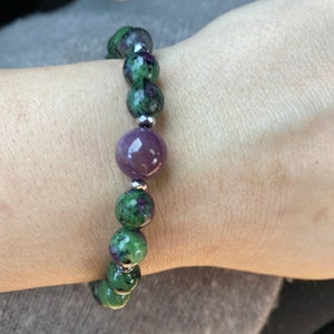 May include: A green and purple gemstone bracelet with a larger purple gemstone in the center. The bracelet is made with silver beads.