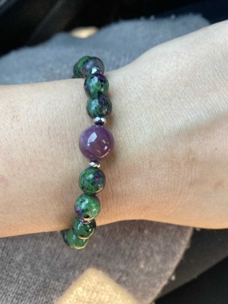 May include: A green and purple gemstone bracelet with a larger purple gemstone in the center. The bracelet is made with silver beads.