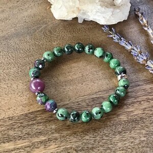 May include: A green and pink gemstone bracelet with silver beads. The bracelet is made of round beads and is on a wooden surface.