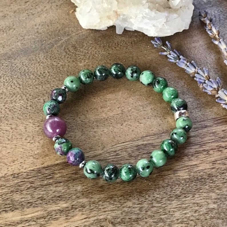 May include: A green and pink gemstone bracelet with silver beads. The bracelet is made of round beads and is on a wooden surface.