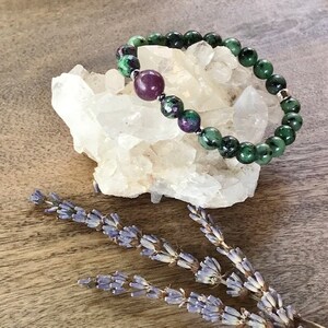 May include: A green and purple gemstone bracelet with a silver clasp. The bracelet is on a white crystal cluster and a sprig of lavender.