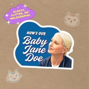 May include: A sticker with a portrait of a woman with blonde hair, the text "HOW'S OUR Baby Jane Doe" in white, and a purple cloud with the text "Glossy, Glitter, or Holographic." Two cat illustrations are also present.