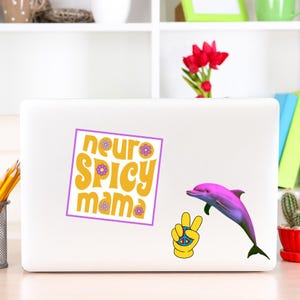 May include: A white laptop with a sticker that says "neuro spicy mama" in yellow and purple. A pink dolphin and a yellow peace sign are also on the laptop. The laptop is on a white desk.