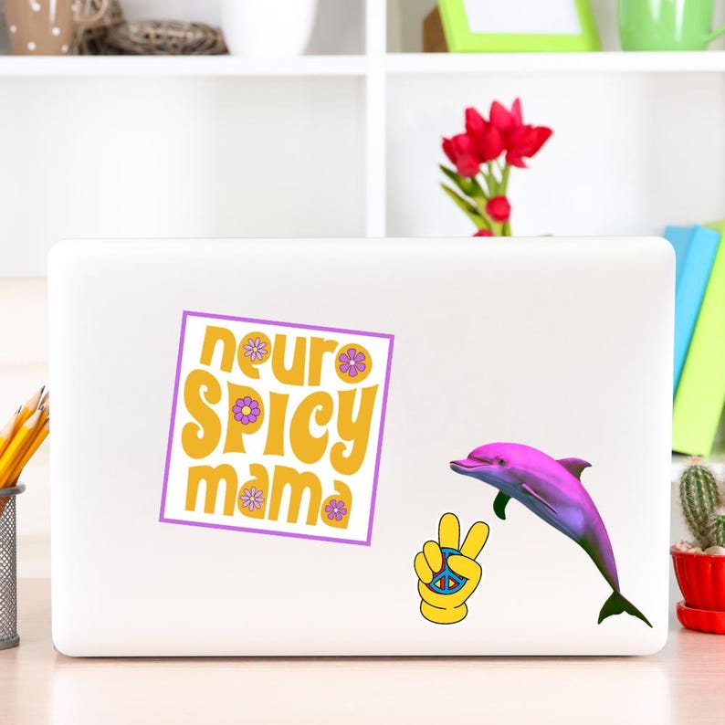 May include: A white laptop with a sticker that says "neuro spicy mama" in yellow and purple. A pink dolphin and a yellow peace sign are also on the laptop. The laptop is on a white desk.