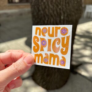 May include: A square sticker with the words "neuro SPICY mama" in orange, with purple flower accents. The sticker is held up in front of a tree trunk.