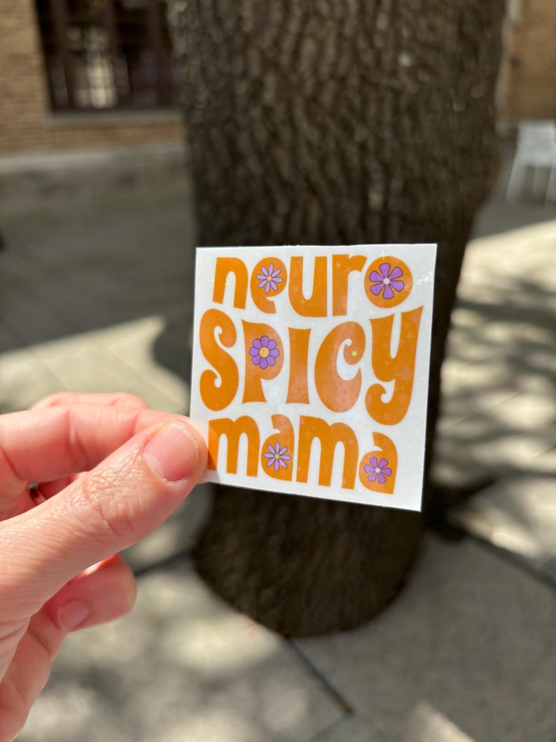 May include: A square sticker with the words "neuro SPICY mama" in orange, with purple flower accents. The sticker is held up in front of a tree trunk.