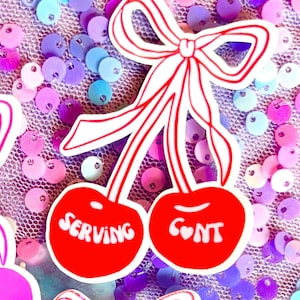 Può includere: Un adesivo a forma di ciliegia rossa con le parole "SERVING" e "C❤NT" in bianco. L'adesivo ha un fiocco bianco con strisce rosse ed è posto su uno sfondo di paillettes colorate.
