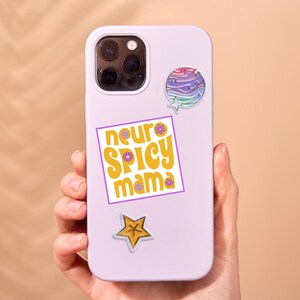 May include: A light purple phone case with multiple stickers. The stickers include a colorful planet design, a star, and a square sticker with the words "neuro spicy mama" in yellow and purple.