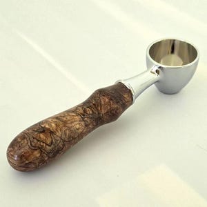 May include: A kitchen scoop featuring a polished silver-colored metal bowl and a cylindrical handle crafted from dark brown wood with a natural, swirling grain pattern. The handle connects to the metal bowl with a smooth, tapered silver ferrule.