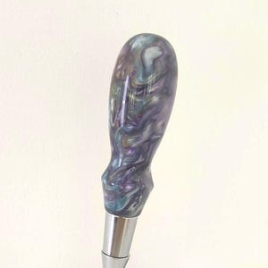 May include: A beer tap handle featuring a polished chrome faucet base and a smooth, teardrop-shaped grip with a swirling, marbled pattern in shades of deep purple, teal, and white. The metal base includes a small, engraved logo and text.