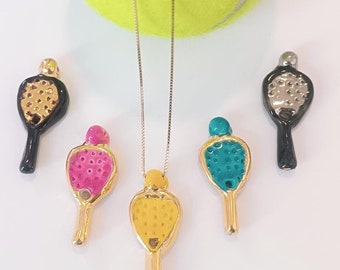 May include: A collection of tennis racket pendant necklaces and charms in various colors, including pink, teal, and yellow. The pendants have gold-colored accents and are displayed with a tennis ball.
