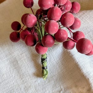 May include: A bundle of faux red berries with a matte finish, tied together with green stems and black wire. The berries are arranged in a cluster and rest on a white linen cloth.