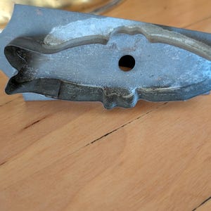 May include: A vintage metal cookie cutter in the shape of a fish. The cutter is gray with a small hole and is resting on a wooden surface. The fish-shaped cutter is a kitchen tool for baking.