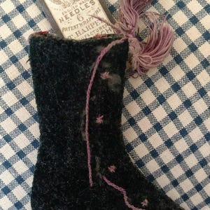 Puede incluir: Un alfiletero en forma de bota de terciopelo negro con bordados rosas y un borla. El alfiletero contiene un paquete vintage de agujas Queen's Crewel.