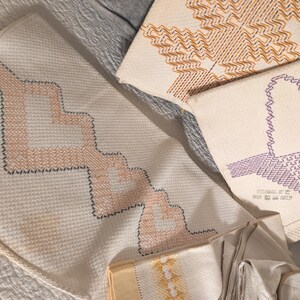 以下が含まれることがあります： クロスステッチ刺繍が施されたオフホワイトの布地3枚。1枚はピーチとダークブルーのジグザグ模様、もう1枚はオレンジの幾何学模様、3枚目はパープルの模様で「STITCHED BY ME MAY 24 2017」という文字が刺繍されています。