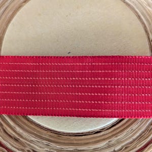 May include: A roll of beige paper with a red ribbon on top. The ribbon is a solid red color with white stitching. The paper roll is tightly wound, with the edges of the paper visible. The image is a close-up shot.