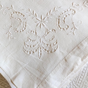 Puede incluir: Servilleta de lino color crema con intrincado diseño bordado floral y de volutas. El bordado presenta un motivo floral central con patrones simétricos y delicados detalles de hojas. Los bordes están cuidadosamente dobladillos con una puntada fina.