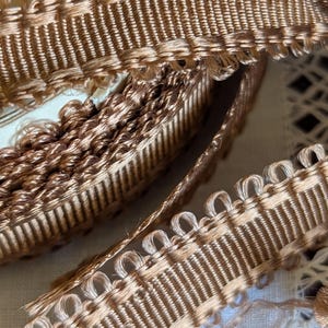 May include: Close-up of tan-colored decorative trim with a looped edge. The trim has a woven texture with vertical lines and a scalloped edge. The trim is likely used for sewing, crafting, or embellishing various items.