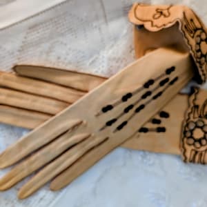 May include: A pair of vintage gloves in a light tan color, with decorative black accents. The gloves have a floral design on the cuffs and a series of black dots on the back of the hand. The gloves are lying on a white lace surface.