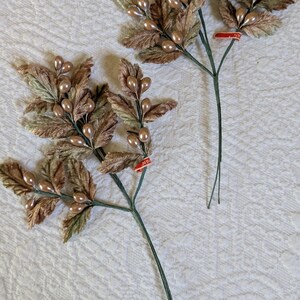 以下が含まれることがあります： 茶色と緑の葉と真珠のような実が付いた、2つの人工的な花の茎。茎は緑色で、葉は質感があります。小さな赤いタグが1つの茎に付いています。背景は白いキルト生地です。