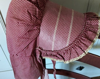 Puede incluir: Un gorro de lunares rojos y blancos con ribete de encaje. El gorro está atado en la parte posterior con una cinta larga.