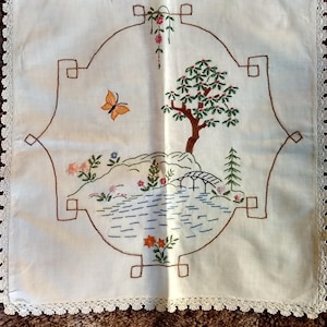 May include: Cream-coloured embroidered square textile with a brown border and a delicate lace trim. The embroidery depicts a landscape with a tree, bridge, butterfly, and flowers in shades of green, orange, and pink.