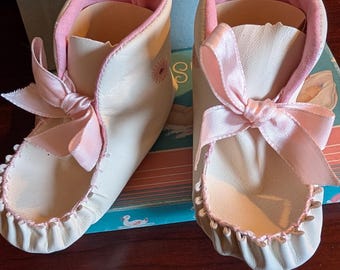 May include: A pair of cream-colored baby booties featuring soft pink satin ribbon laces, pink interior lining, and a small embroidered flower detail on the side. The booties are presented in a light blue cardboard box with a swan illustration.
